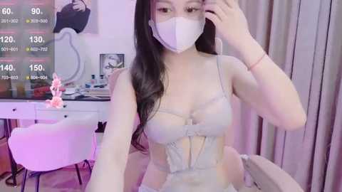 Media: A video of an East Asian woman in a sheer, beige lace lingerie set, wearing a face mask, with long dark hair, seated in a modern, softly lit room.