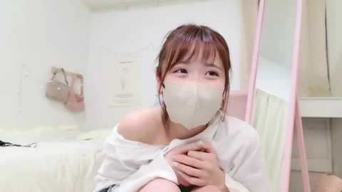 Media: A young Asian woman with short brown hair, wearing a white mask and off-shoulder white shirt, sits on a bed in a minimalist room with white walls and a pink mirror.