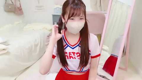 Media: Video of an East Asian woman with long brown hair in a white cheerleader outfit, mask, and red shorts, sitting on a floor in a pink room with a bed, mirror, and white curtains.