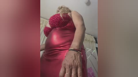 Video of a curvy, blonde woman in a tight, shiny red dress with sequins, wearing a bracelet, leaning forward in a bedroom with beige curtains and a white bed frame.