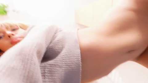 Video of a light-skinned woman with blonde hair, wearing a gray sweater, exposing her bare, slim waist and flat stomach, taken from a low angle, indoors with blurred background.