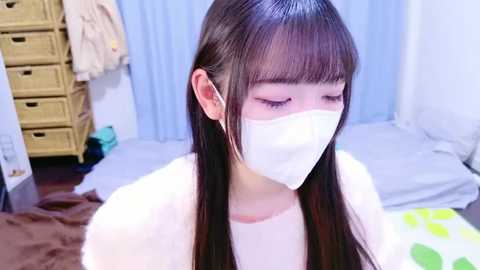 Media: Video of a young East Asian woman with straight, dark hair, wearing a white mask and light-colored top, sitting in a cluttered bedroom with wicker storage and a bed.