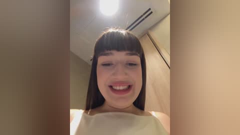 Media: Video of a young Caucasian woman with straight brown hair and blunt bangs, smiling widely. She wears a white tank top. Background shows beige walls and a ceiling light.