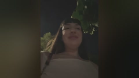 Media: A low-quality video of a young Asian woman with long black hair, wearing a white shirt, standing in a dark, leafy outdoor setting. Her expression is neutral.