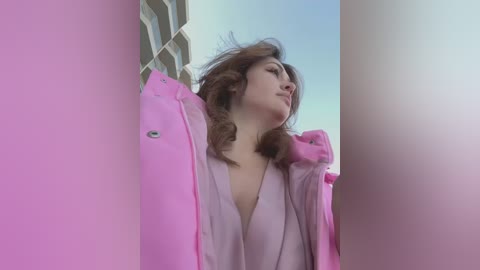 Media: Video of a young woman with wavy brown hair, wearing a pink jacket, standing between two buildings under a clear sky.