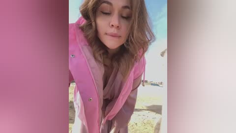 Media: Video of a fair-skinned woman with wavy brown hair, wearing a pink raincoat, smiling softly, standing outdoors on a sunny day.