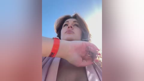 Media: Video of a young woman with light brown hair, wearing a red wristband and a light-colored top, looking upward against a clear blue sky.