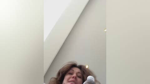 Media: A video of a woman with shoulder-length brown hair, captured from a low angle, showing her from the waist up. The background features a minimalist, modern interior with off-white walls and a slanted ceiling.