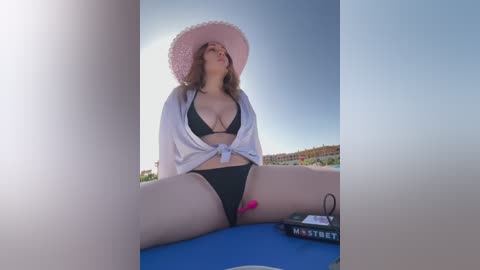 Media: Video of a curvy, fair-skinned woman with medium-length brown hair, wearing a pink sun hat, black bikini, and white robe, sitting on a blue mat outdoors.