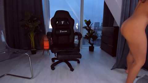 Media: Video of a modern, dimly lit office with a large window, showcasing a black gaming chair, plants, and a partially visible nude person.