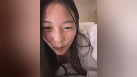 Media: A close-up video of a young Asian woman with long, straight black hair, wearing a white shirt, smiling playfully. The background shows a white bed with a pillow.