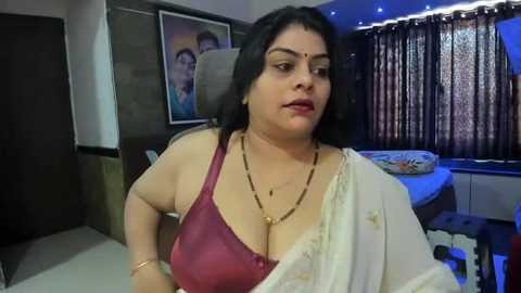 Media: Video of a plus-sized South Asian woman in a maroon bra and white sari, wearing a gold necklace. Background features a dark room with a framed photo and curtains.