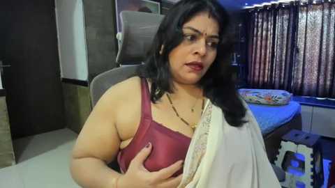 Media: Video of a plus-sized South Asian woman with dark hair, wearing a red bra, and a beige sari, seated in a modern bedroom with a bed and curtains in the background.