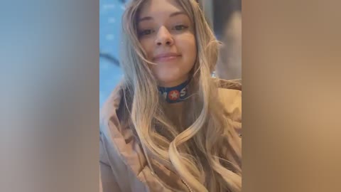 Video of a young woman with long, wavy blonde hair, wearing a beige coat and a blue choker with a cartoon character design. She has fair skin and is smiling slightly. Background is out of focus.