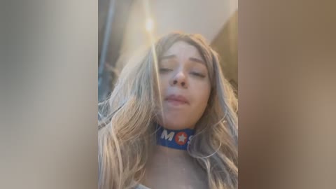 Media: Video of a young woman with long, wavy blonde hair, wearing a blue choker with \"M&M's\" logo, smiling with closed eyes, surrounded by soft, warm lighting, creating a dreamy, slightly blurry effect.