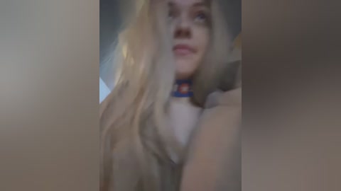 A blurred video of a blonde woman with long hair, wearing a blue collar, and a black top, taken indoors with a soft, muted lighting.