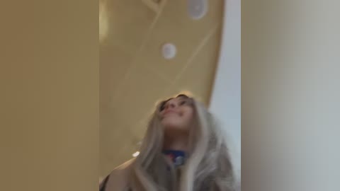 A video of a woman with long blonde hair, wearing a beige coat, standing indoors against a beige wall. The image is slightly blurred, with a neutral background.