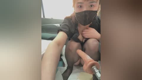 Media: Video of an Asian woman with fair skin, wearing a black mask, black dress, and sheer stockings, sitting on a car seat with a dildo inserted into her vagina.