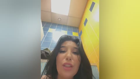 Media: A video of a young woman with long, dark hair, wearing a yellow shirt, in a brightly lit bathroom with blue and yellow tiled walls. She has a neutral expression.