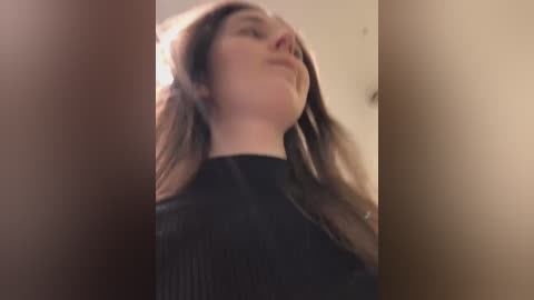 Media: A video captures a young woman with long brown hair, wearing a black ribbed sweater, looking upwards, in a dimly lit, beige room.