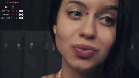 Media: Video of a close-up of a woman with light brown skin, wearing dark eyeliner and a nose ring, against a blurred, dark background.