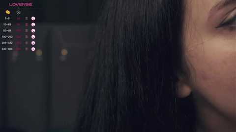 Media: Video of a close-up of a person's face with long, straight black hair, showing the ear and side profile. The background is blurry and dark, with a digital interface displaying \"Lovingly\" and various chat options.