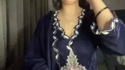 Media: Video of a woman with medium skin tone wearing a dark blue velvet dress with intricate white embroidery and a pink floral design. She has shoulder-length dark hair and a silver necklace. Background features neutral-toned curtains and a dimly lit room.