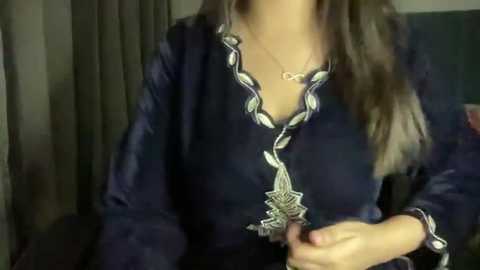 Media: Video of a woman with long, wavy brown hair wearing a dark blue satin robe with intricate silver embroidery and a matching necklace. She sits against a dark curtain backdrop, her hands resting on her lap.