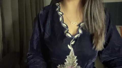 Media: Video of a woman wearing a dark velvet blouse with a silver necklace featuring an intricate, geometric pendant. She has long, straight, brown hair. The background includes a dark curtain.