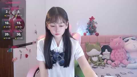 Media: Video of an Asian woman with long black hair, wearing a white blouse and plaid bow, sitting in a room with plush toys, a pink bed, and a Christmas tree.