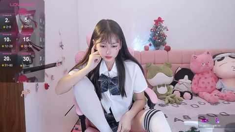 Media: Video of a young East Asian woman with long black hair, dressed in a schoolgirl outfit, sitting on a pink chair in a cozy room with a decorated Christmas tree and plush toys.