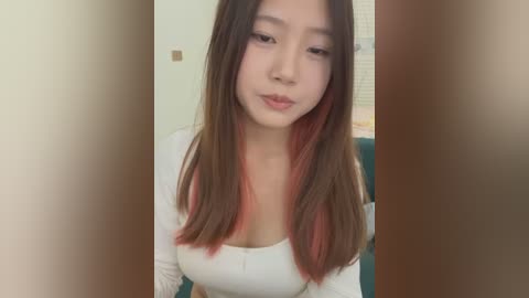 Media: Video of an East Asian woman with straight, shoulder-length hair, fair skin, and a white long-sleeve top. She has a neutral expression and is seated indoors with a blurred background.