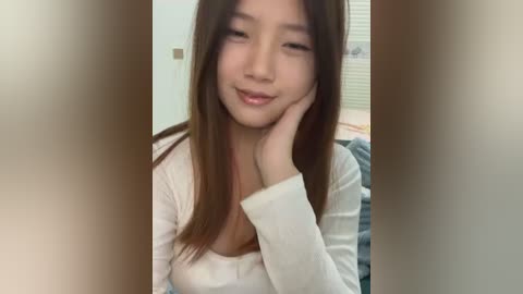 Media: Video of a young Asian woman with straight, long brown hair, light skin, wearing a white long-sleeved top, leaning on her right hand, smiling softly.