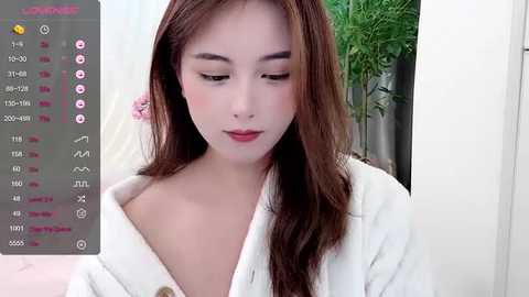 Media: A video of a young Asian woman with fair skin and long brown hair, wearing a white robe, looking down with a serene expression. The background features a green plant and soft lighting.