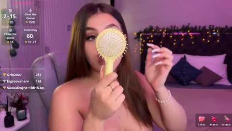 Media: A topless young woman with long brown hair, holding a yellow hairbrush close to her face, in a bedroom with a bed, Christmas lights, and makeup on a table.