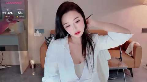 Video of an East Asian woman with long black hair, red lipstick, wearing a white blouse and black top, adjusting her hair in a modern living room with a sofa and a digital clock displaying 21:66.