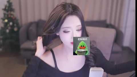 Media: Video of an Asian woman with long black hair, wearing a black off-shoulder top, taking a selfie. Background includes a grey sofa, a Christmas tree, and a TV screen showing a green Angry Bird character.