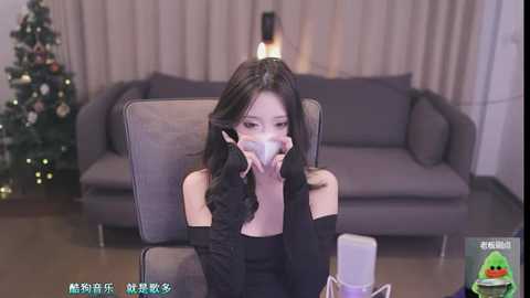 Media: A video of an Asian woman with long black hair, wearing a black off-shoulder top, sitting on a gray couch, holding a white mask, with a Christmas tree in the background.