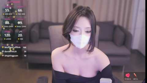 Media: Video of an Asian woman with short black hair, wearing a white face mask and black off-shoulder top, sitting on a couch. Background includes a gray sofa and dim lighting.