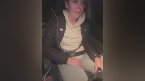 Media: Video of a woman with short dark hair, wearing a beige hoodie and gray pants, driving a car at night, with a dimly lit interior and blurred background.