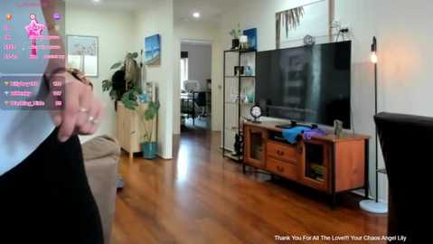 Media: Video of a modern living room with a wooden floor, a large flat-screen TV on a wooden cabinet, a person's hand holding a controller, and a green plant in the background.