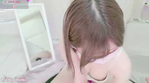 Media: A video of an Asian woman with straight, shoulder-length brown hair, wearing a white bra, kneeling on a tiled floor. She is in a bathroom with a mirror and a white sink visible in the background.