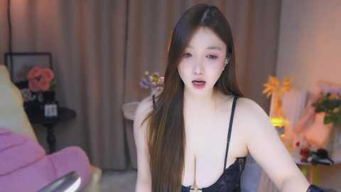 Media: Video of a fair-skinned, long-haired East Asian woman in black lingerie, sitting on a chair, indoors with a pink couch, flowers, and a lamp in the background.