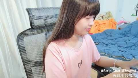 Media: Video of an East Asian girl with long brown hair in a pink T-shirt, sitting on a black mesh chair, in a messy bedroom with blue bedding and orange clothes.