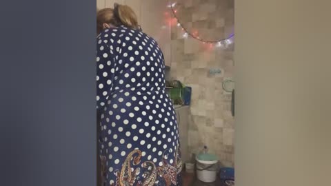 Media: Video of a woman in a navy blue polka-dot dress bending over, with a colorful patterned bag in hand, standing in a tiled bathroom with string lights and toiletries.