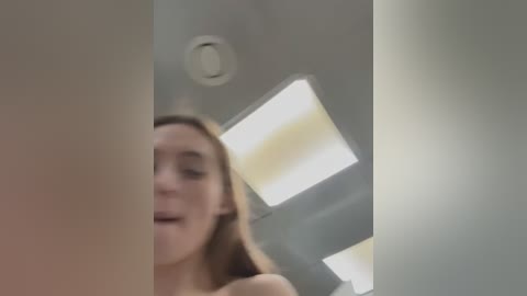 Media: Video of a young woman with long brown hair, wearing a strapless top, standing in a modern room with a large skylight and white walls. The image is slightly blurred.