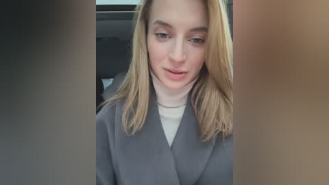 Media: Video of a blonde woman with fair skin and a white turtleneck, wearing a gray coat, seated in a car. The background shows the car's interior and blurred figures.