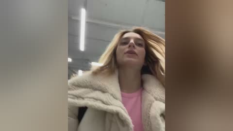 Media: Video of a Caucasian woman with long, blonde hair, wearing a pink top under a beige fur coat, captured from a low angle in a dimly lit, industrial setting with visible fluorescent lights.