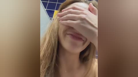 Media: Video of a smiling woman with long blonde hair, partially covering her face with her hand. She is in a restroom with a blue and white tiled ceiling.