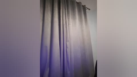 Media: Video of a plain, off-white wall with a long, sheer, gray curtain hanging vertically, slightly pulled to the right, revealing a soft, diffused light source behind it.